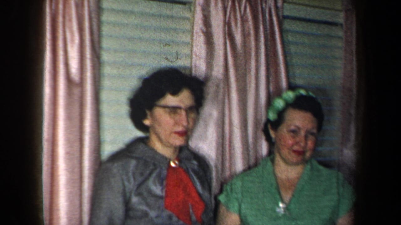 Two women stand together inside a New York home, one in a gray dress with red neck detail and one in a green dress.
