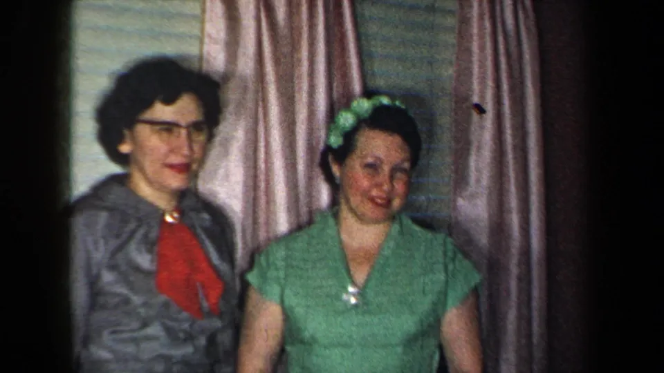 The two women remain posed in the private interior, framed closely against curtains and blinds in the 1959 home movie.