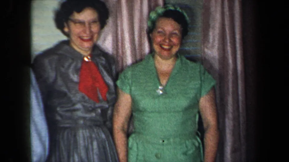 Two women smile toward the camera in a 1959 New York home interior, with curtains and blinds visible behind them.