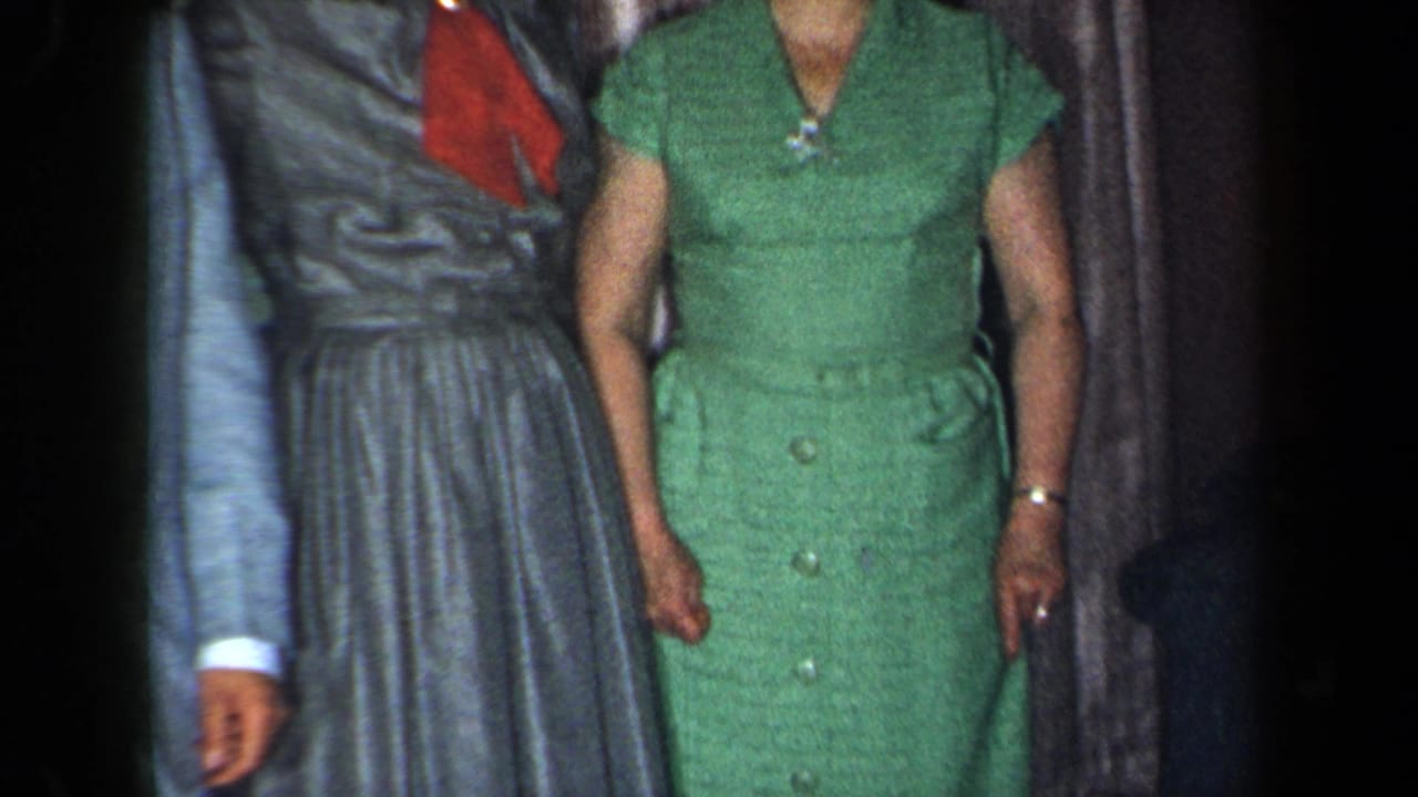 Adults in formal clothing stand indoors, cropped at the faces, with a green dress and grey skirt visible.