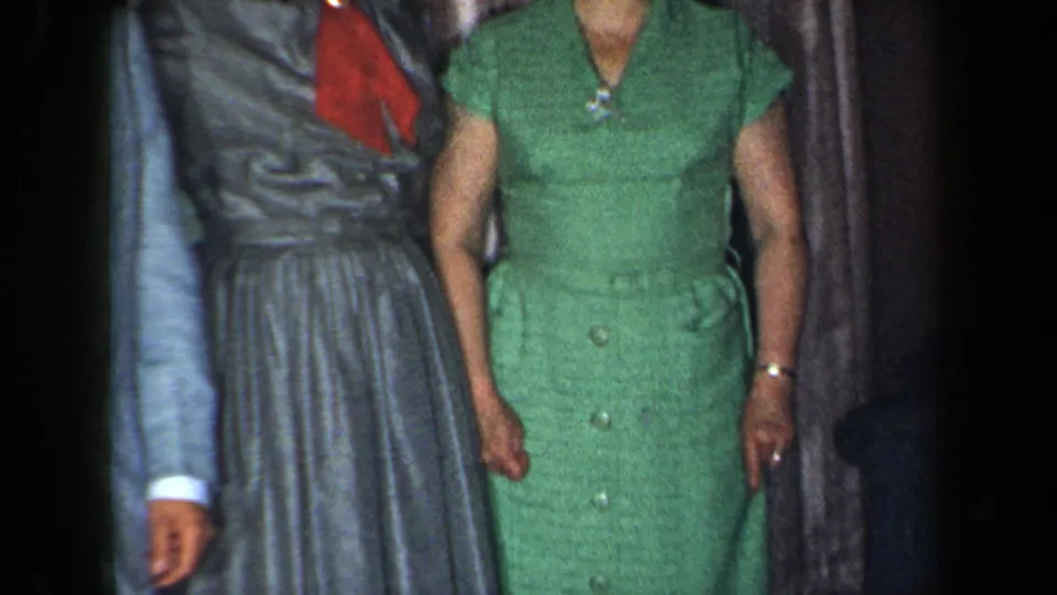 1959 New York home-movie frame showing adults posed indoors in a light blue suit, grey dress, and green button-front dress.
