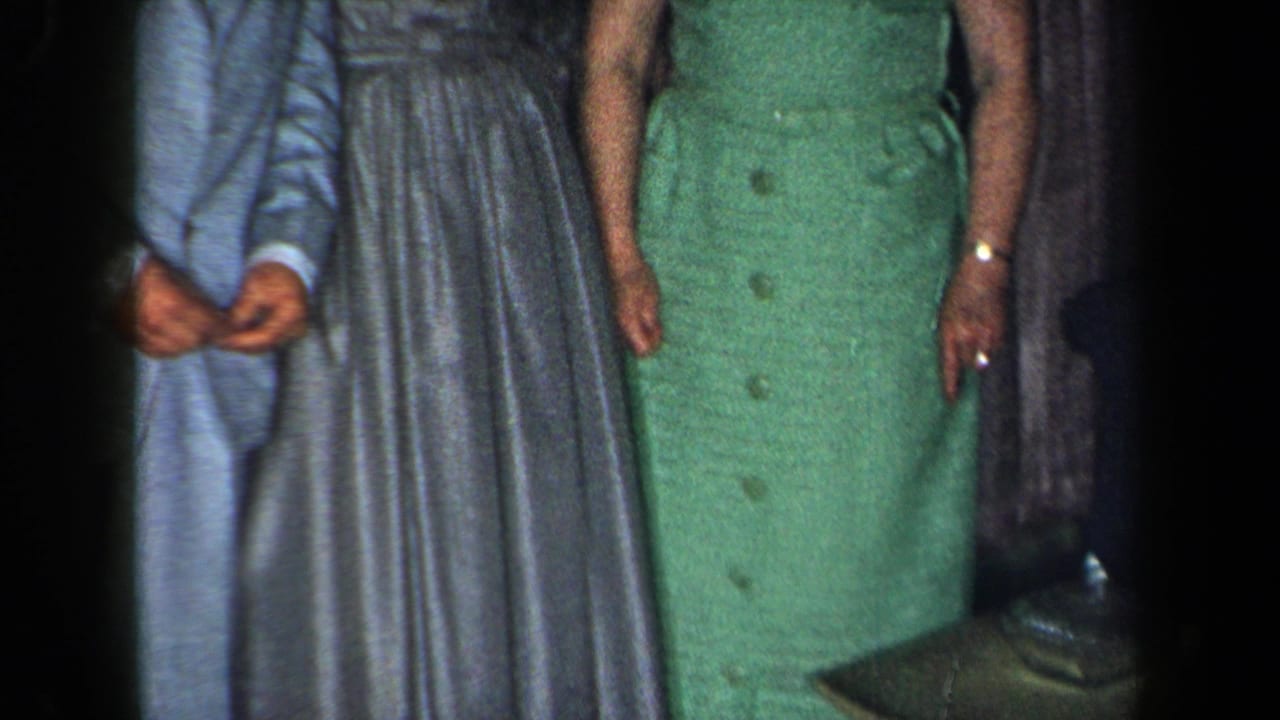 1959 New York home-movie frame with a man in a light blue suit beside women in grey and green dresses indoors.
