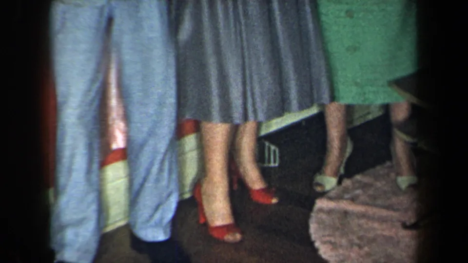 1959 New York home-movie frame showing cropped figures indoors, with a light blue suit, grey dress, green dress, and red heels.