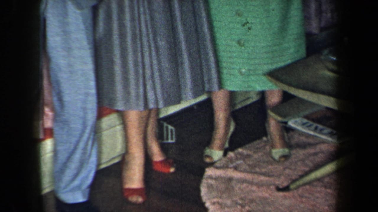 Cropped view of adults standing indoors near a coffee table, showing dresses, high heels, and part of a light suit.