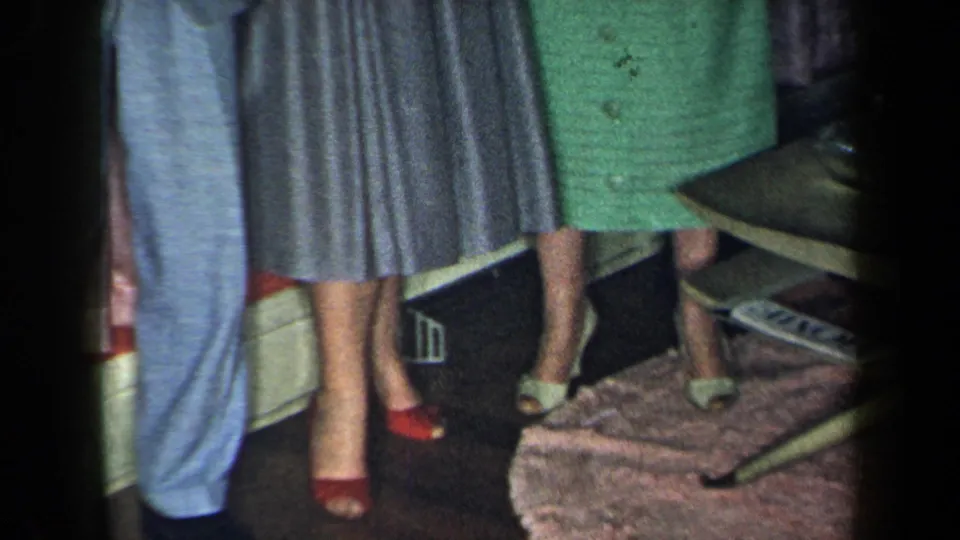 1959 New York home-movie frame with cropped adults in dress clothes, including red and white heels, a green dress, curtains, and a coffee table.