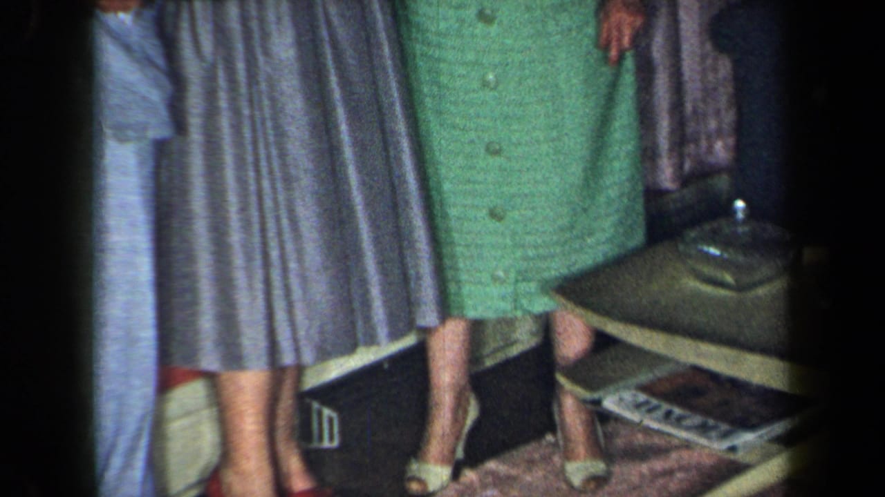 1959 New York home-movie frame showing adults in dress clothes, a green dress, gray skirt, high heels, coffee table, magazines, and glass ashtray.