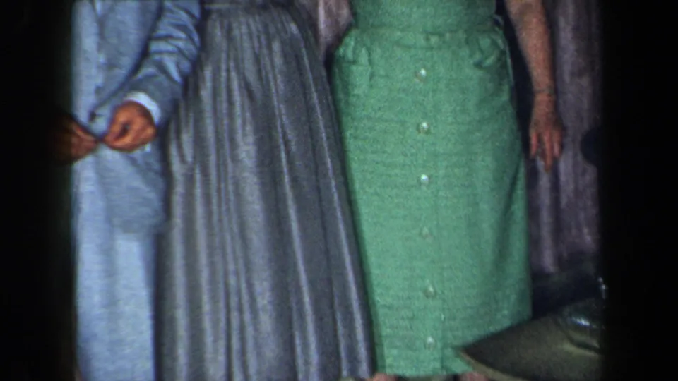 1959 New York home-movie frame with adults standing together indoors, including a light suit, gray pleated dress, green dress, curtains, and coffee table edge.