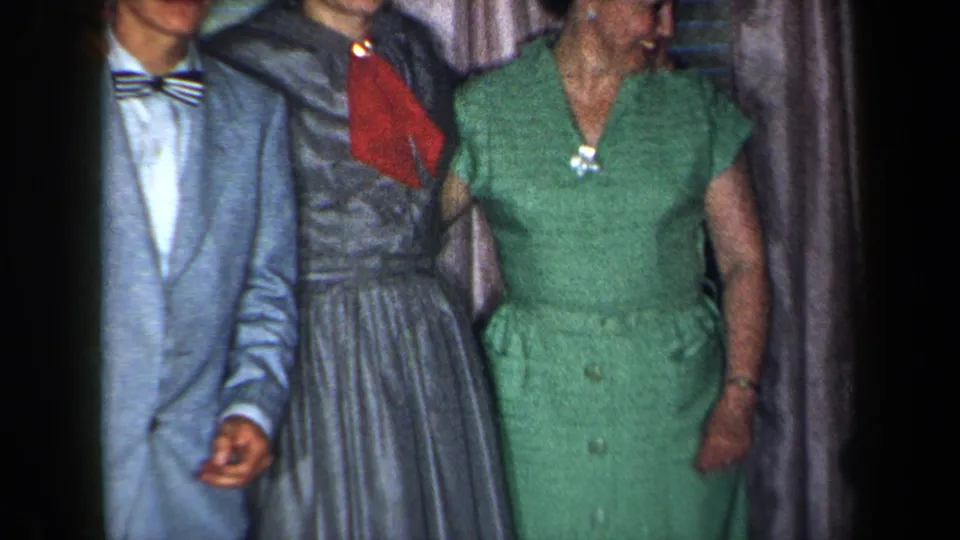 1959 New York home-movie frame showing a man in a light suit and bow tie beside women in gray and green dresses, with faces partly cropped and curtains behind.
