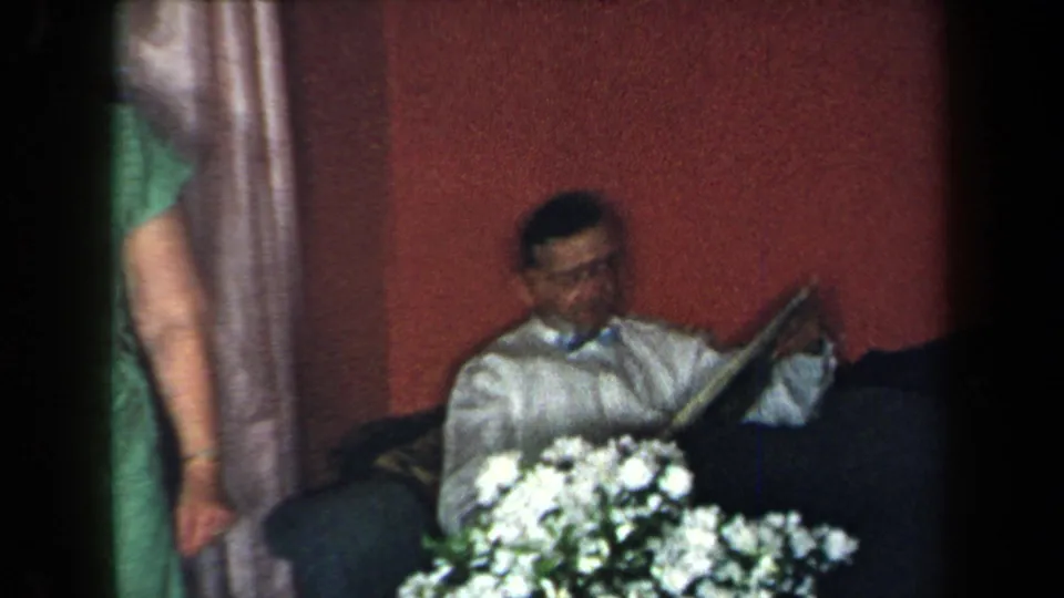 1959 New York home-movie frame of a seated man reading a newspaper in a red-walled room as a woman in green stands nearby.