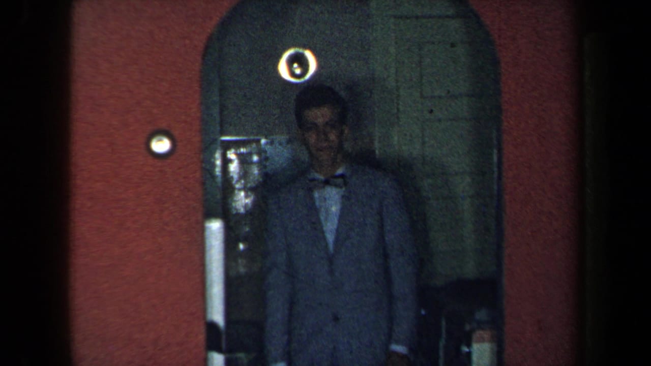 Man in a suit and bow tie stands in an arched doorway against red walls.