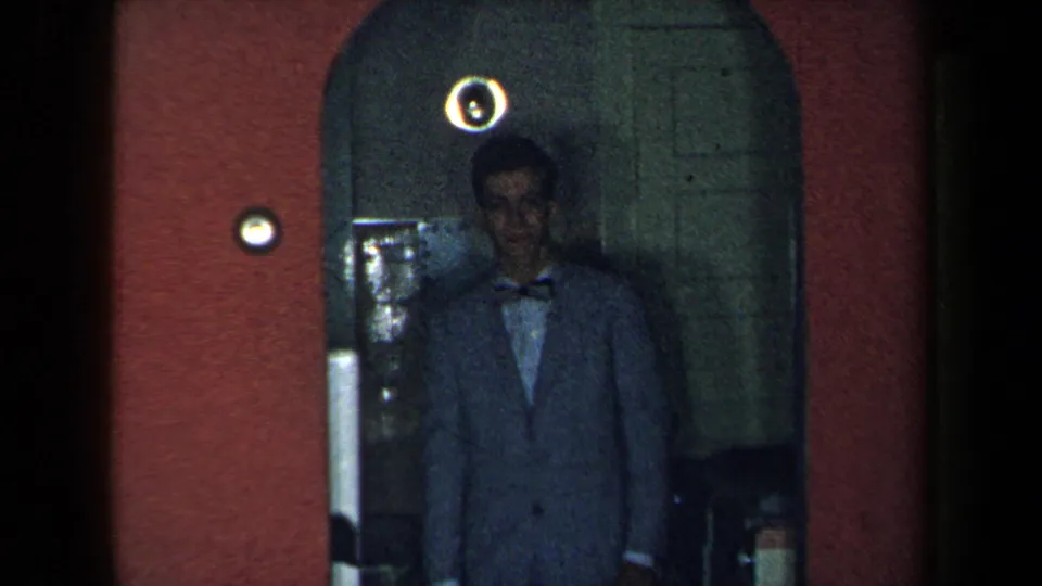 1959 New York home-movie frame showing a man in a light suit and bow tie standing in an arched interior doorway.