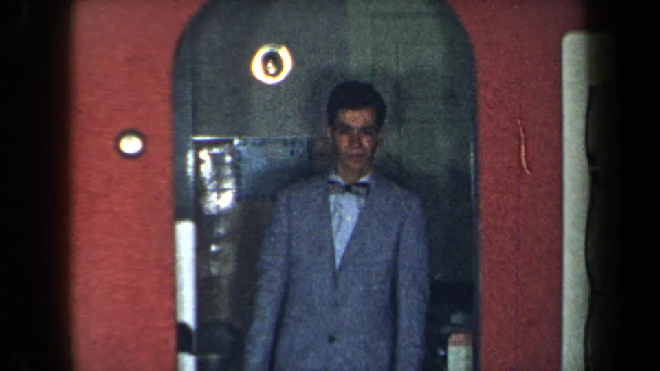 1959 New York home-movie frame of a man in a light blue suit and bow tie standing frontally in a red-walled doorway.