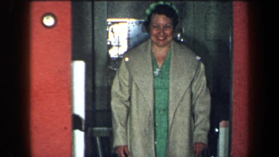 1959 New York home-movie frame of a smiling woman in a beige coat and green dress entering or standing in an interior doorway.