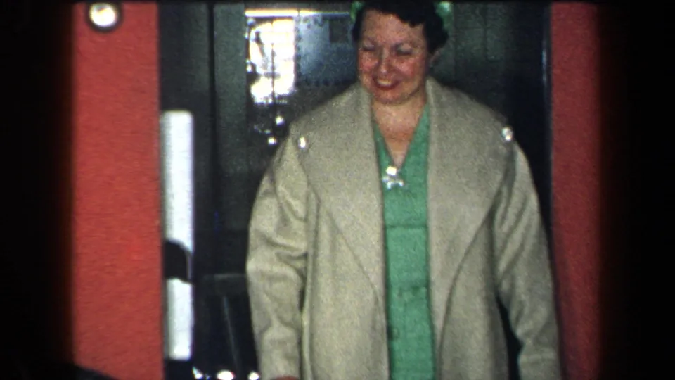 In 1959 New York home-movie footage, a woman in a beige coat and green dress smiles while stepping through an interior doorway.