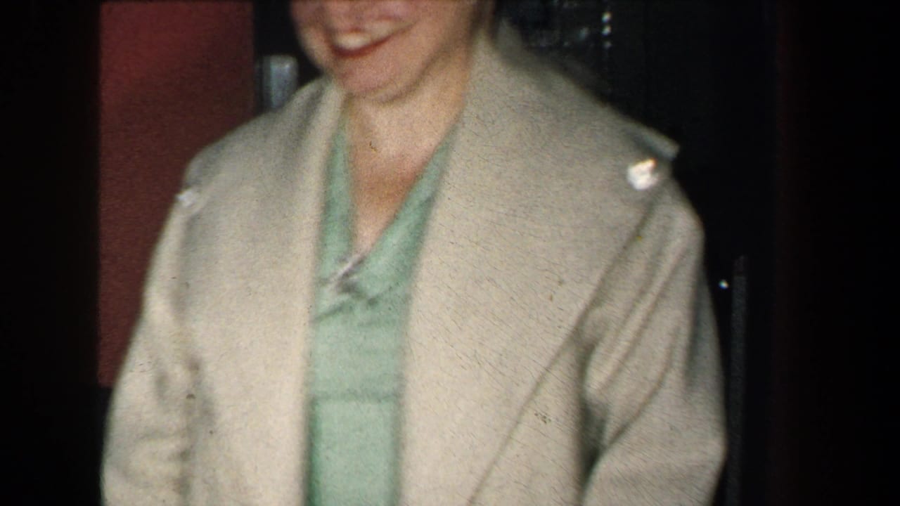 A closer 1959 New York frame shows the woman smiling in a beige coat and green dress beside a dark interior doorway.