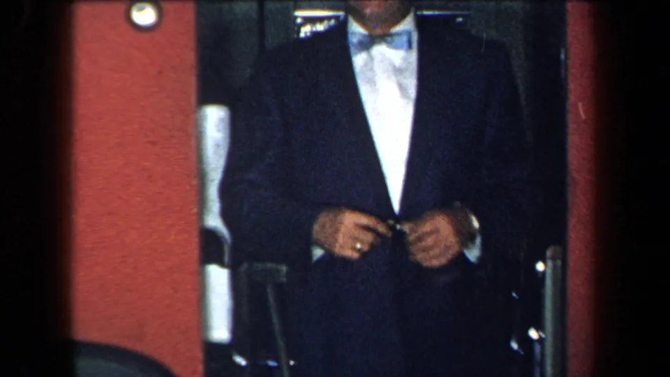 A man in a dark suit, white shirt, and bow tie appears partly cropped as he enters the same dim 1959 New York interior.