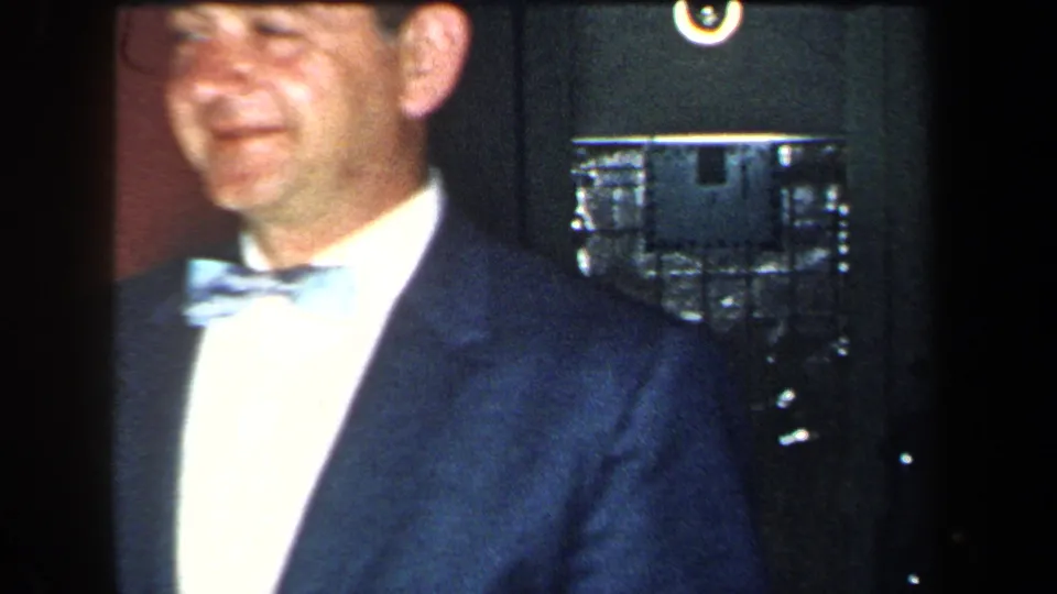 A close 1959 New York home-movie frame shows a smiling man in a dark suit and pale bow tie inside a private room.