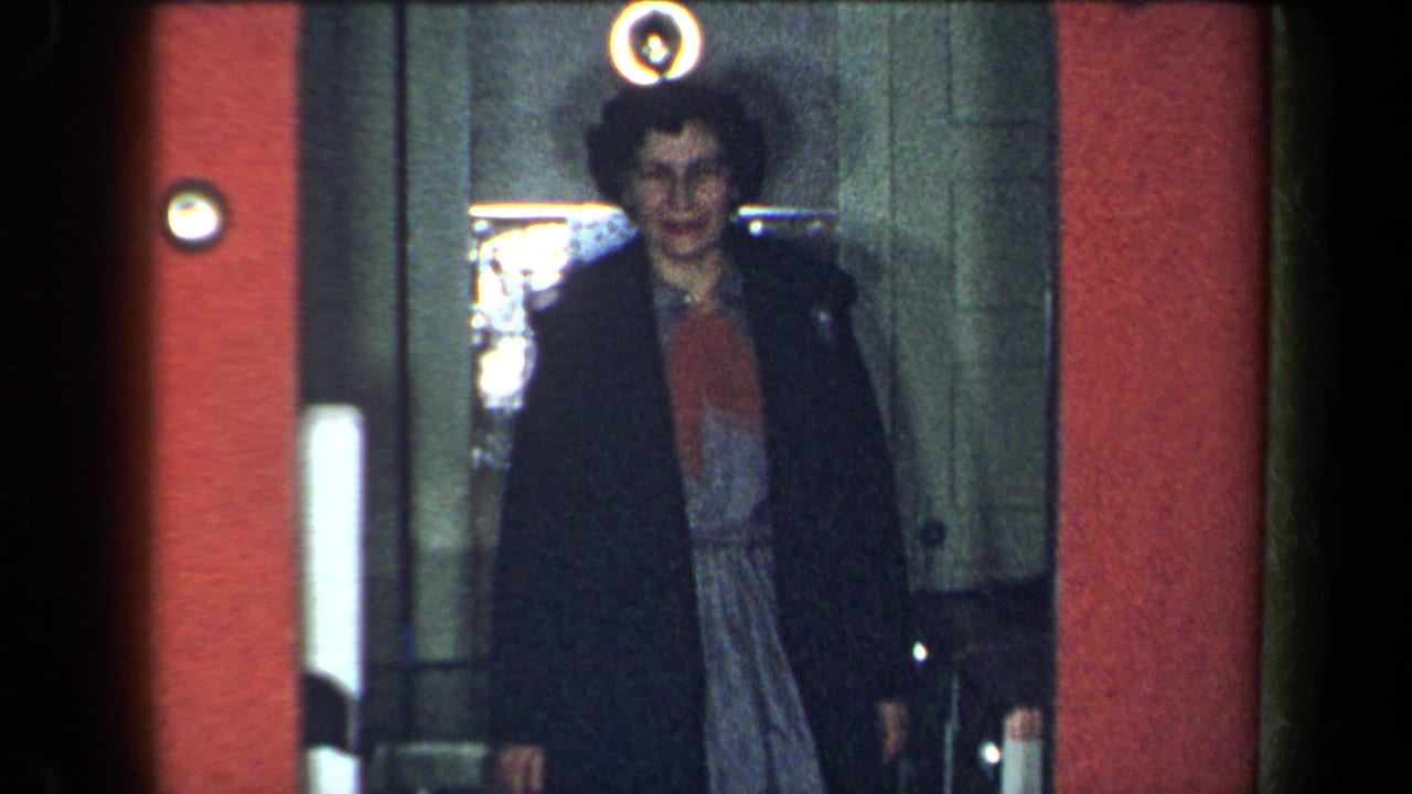 1959 New York home-movie frame of a woman emerging from an elevator, smiling, with a red wall and round light nearby.