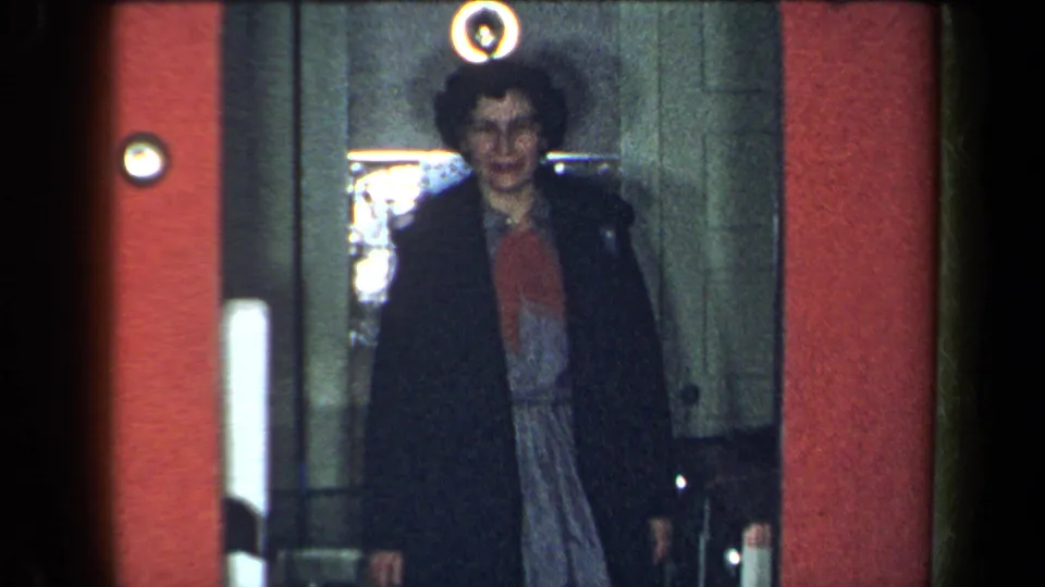 1959 New York home-movie frame of a woman emerging from an elevator, smiling, with a red wall and round light nearby.