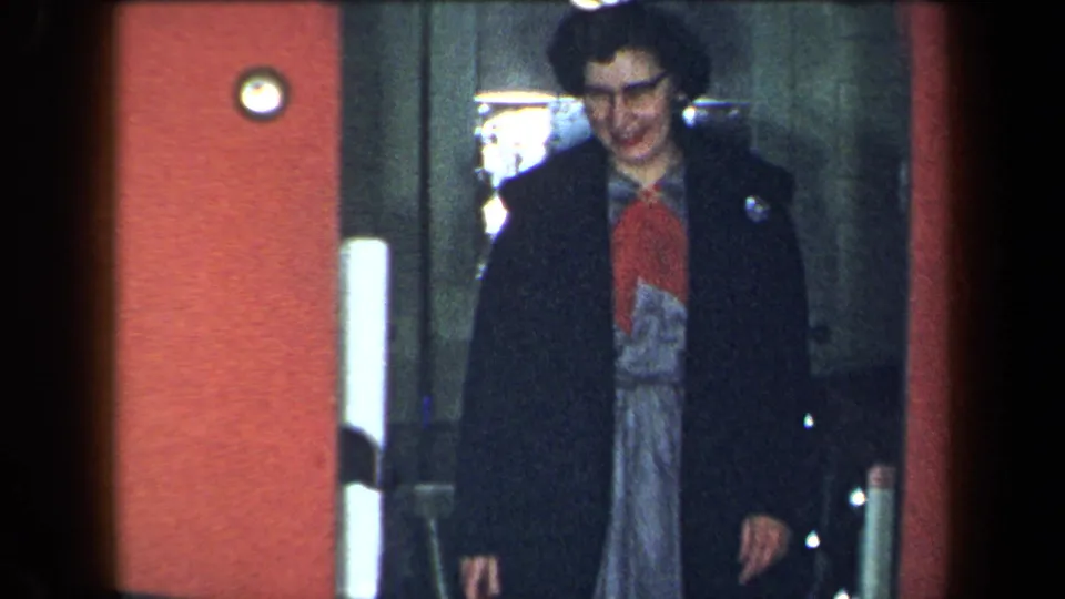 1959 New York 8mm frame showing the woman in a dark coat and patterned dress stepping forward from the elevator.