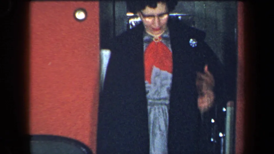 1959 New York home-movie frame with a closer view of the woman’s coat, patterned dress, red wall, and elevator doorway.