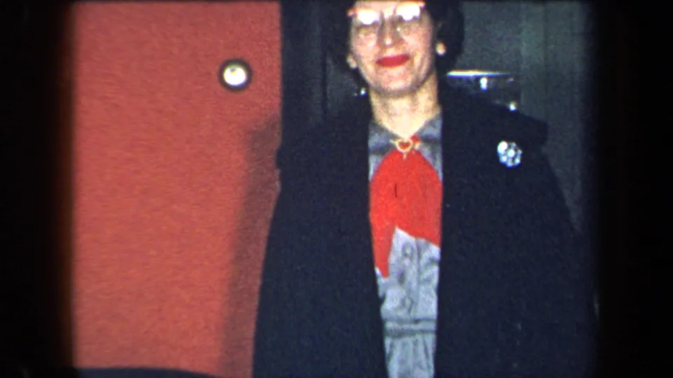 In a 1959 New York home-movie frame, a woman in glasses, red scarf and dark coat faces the camera near a red wall and doorway.