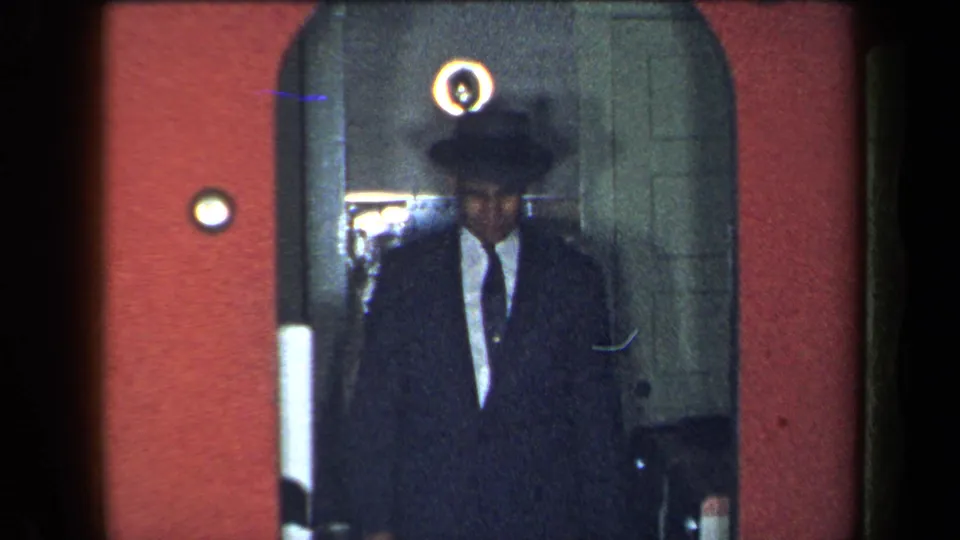 The suited man in a brimmed hat remains in the interior doorway, framed by the red wall and glass door in the 1959 New York home movie.