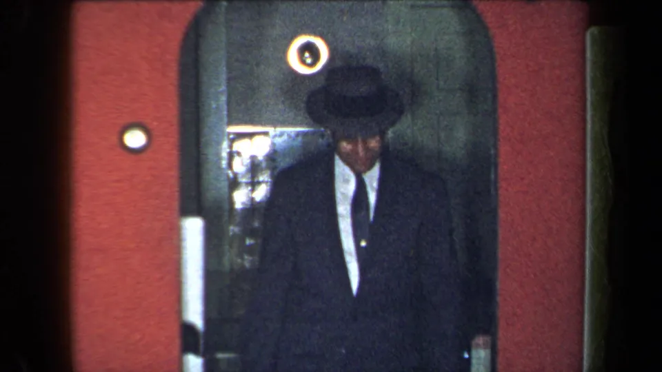 In 1959 New York home-movie footage, a suited man in a dark brimmed hat appears in shadow inside an arched doorway.