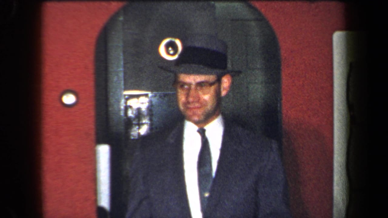 Man in suit and brimmed hat facing the camera near a red wall and arched doorway.