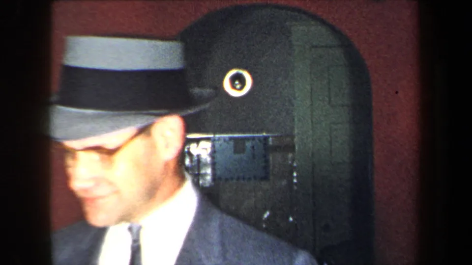 A close, slightly overexposed view shows the suited man in a gray-and-dark brimmed hat passing near the camera.
