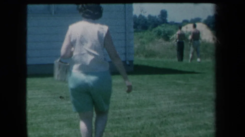 A woman in light clothing carries a basket across a lawn, with two distant men near a house in 1959 New York home-movie footage.