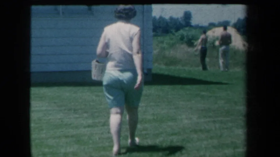 A woman walks away across a sunny lawn carrying a basket, while two men remain in the background near a house.