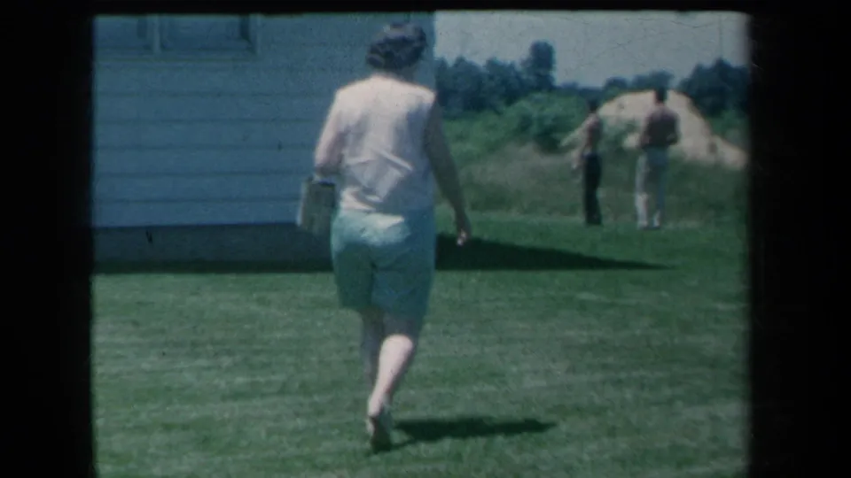 In a 1959 New York home-movie frame, a barefoot woman in a light sleeveless top and green shorts walks across a lawn toward two men in the distance.