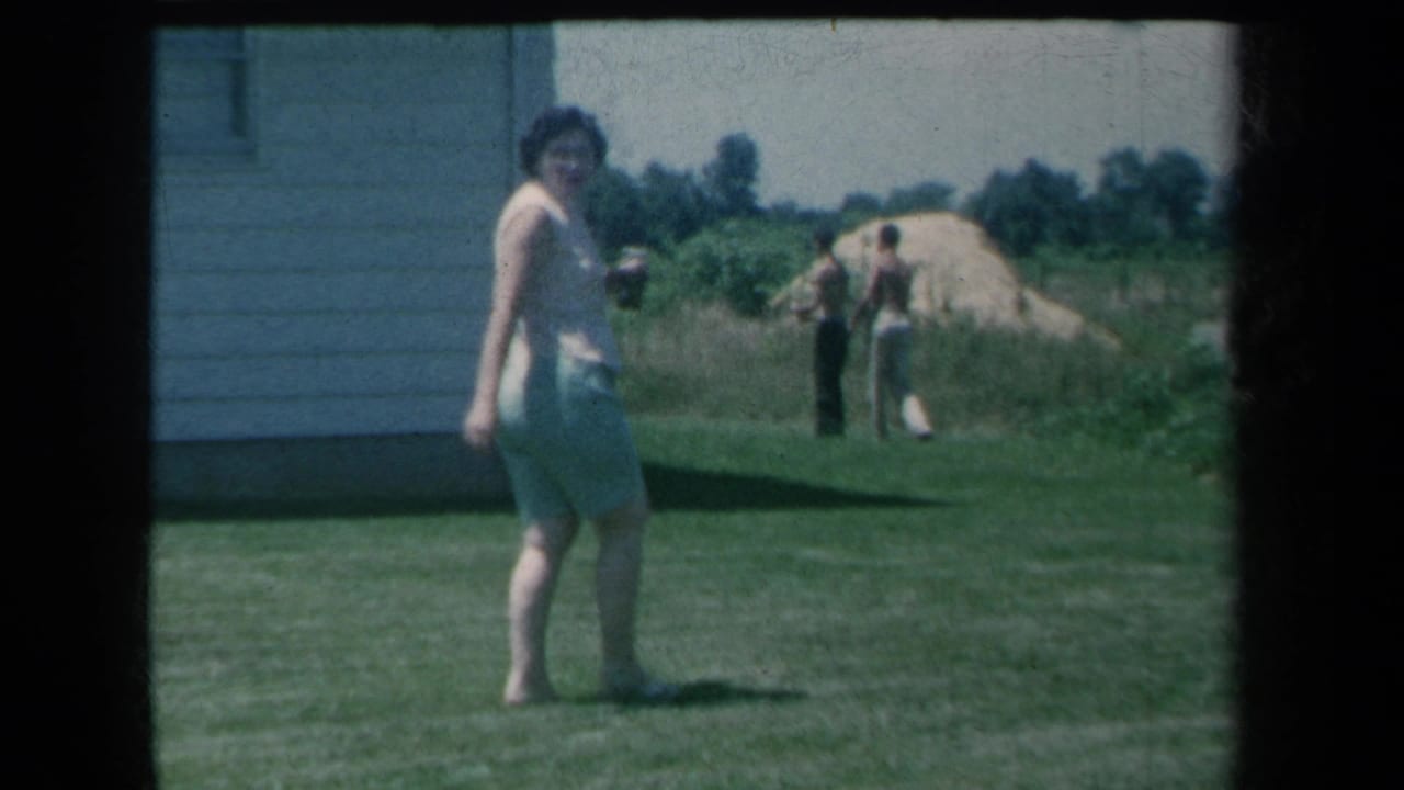The woman turns back toward the camera in the yard, with two men, sand piles, trees, and the side of a house visible behind her.