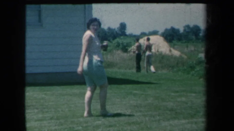 The woman turns back toward the camera in the yard, with two men, sand piles, trees, and the side of a house visible behind her.