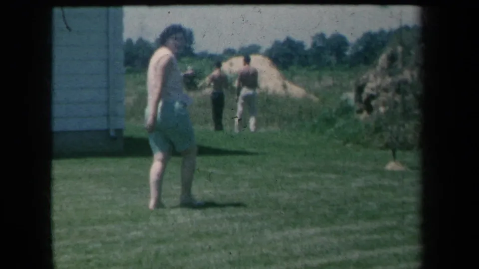 A woman pauses or turns on the grass, facing the camera while two men stand near sand piles beyond the side of a house.