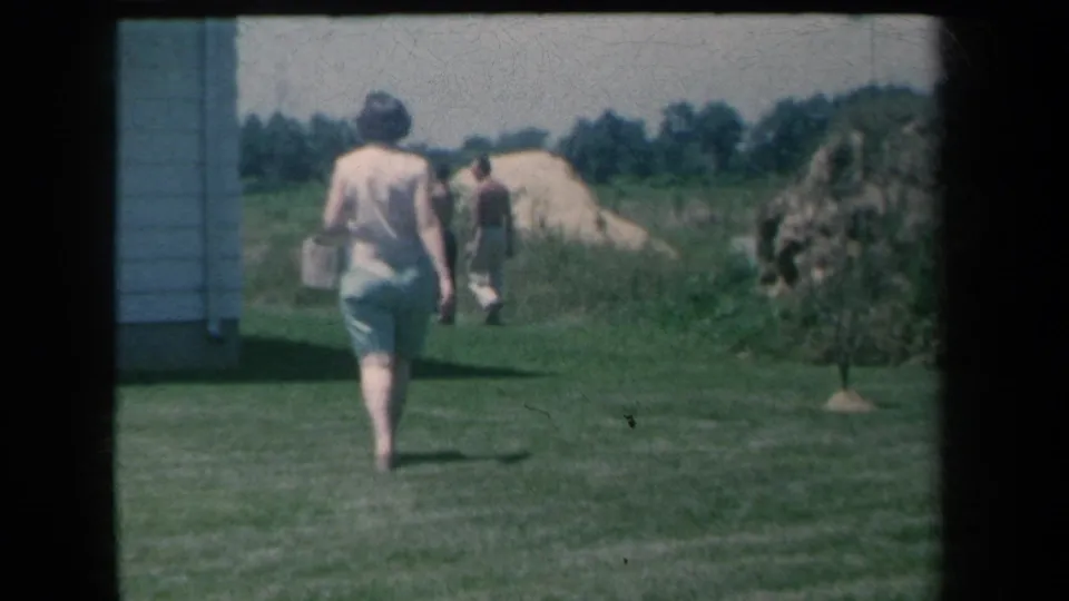 The woman continues away from the camera across the New York yard, carrying a small bag as two men stand near sand piles and trees.