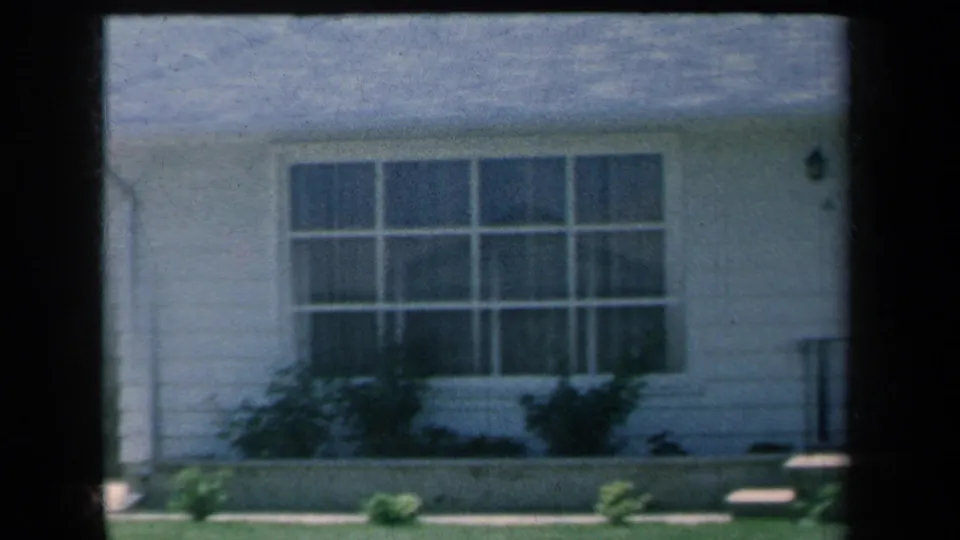 1959 8mm frame of a white suburban New York house facade with a broad front window, low shrubs, lawn, and visible film gate edges.
