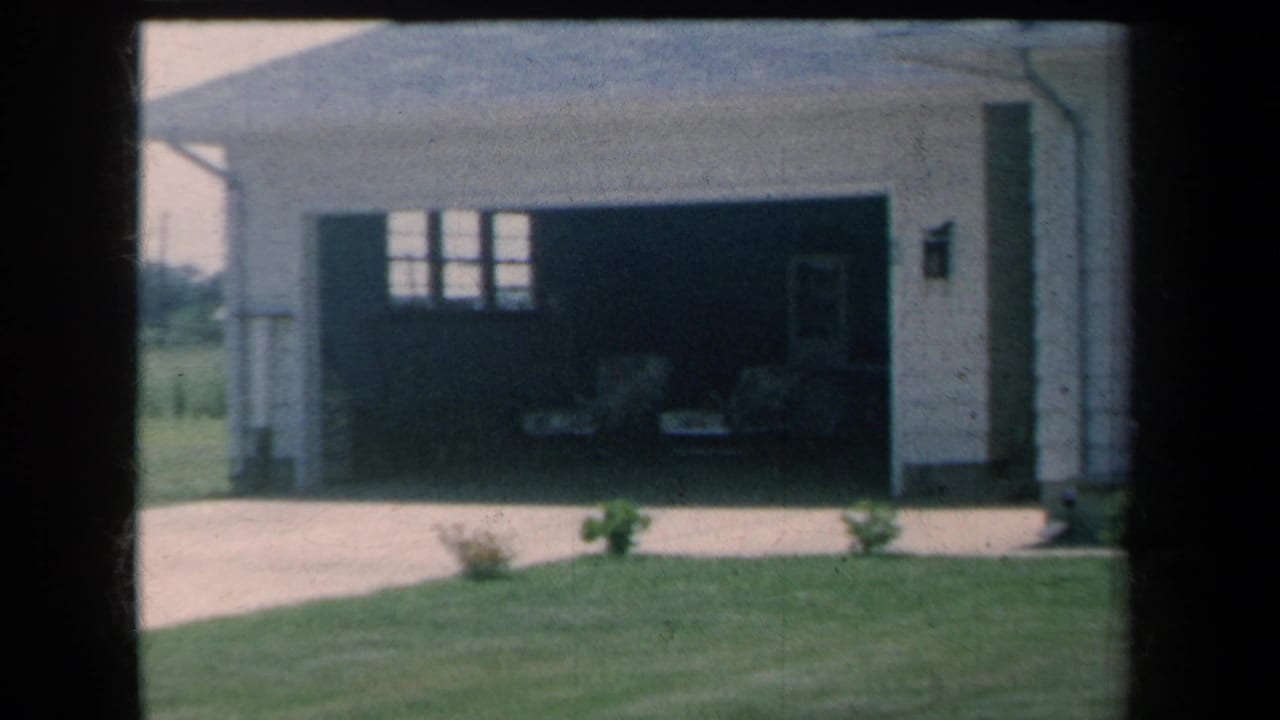 1959 8mm view of a suburban New York house garage standing open beside a driveway, lawn, small shrubs, and pale exterior siding.