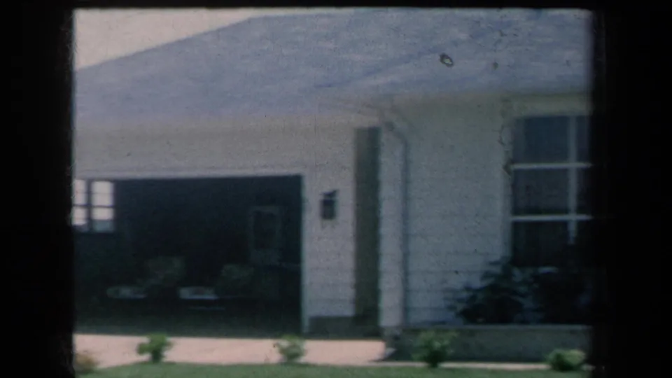 1959 8mm frame showing the house corner between an open garage and front window, with driveway, shrubs, and lawn in view.