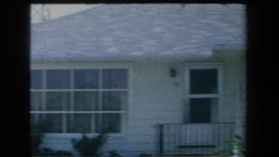 1959 8mm view of a suburban New York house facade with a front door, porch railing, large multi-pane window, shrubs, and lawn.