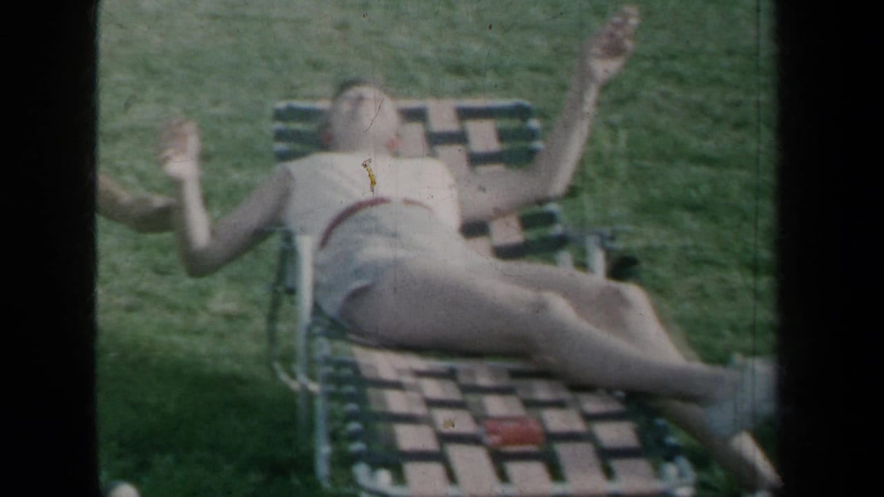 1959 New York home-movie frame showing a woman reclining on a lawn chair and a shirtless man seated nearby on grass.