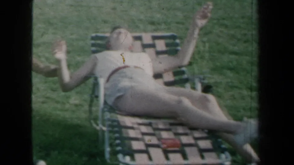 1959 New York home-movie frame showing a woman reclining on a lawn chair and a shirtless man seated nearby on grass.