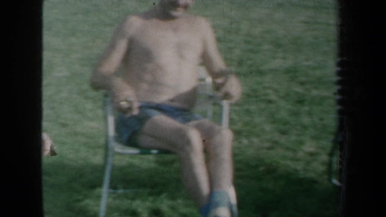 1959 New York frame of a shirtless man in swim trunks sitting in a folding lawn chair on grass.