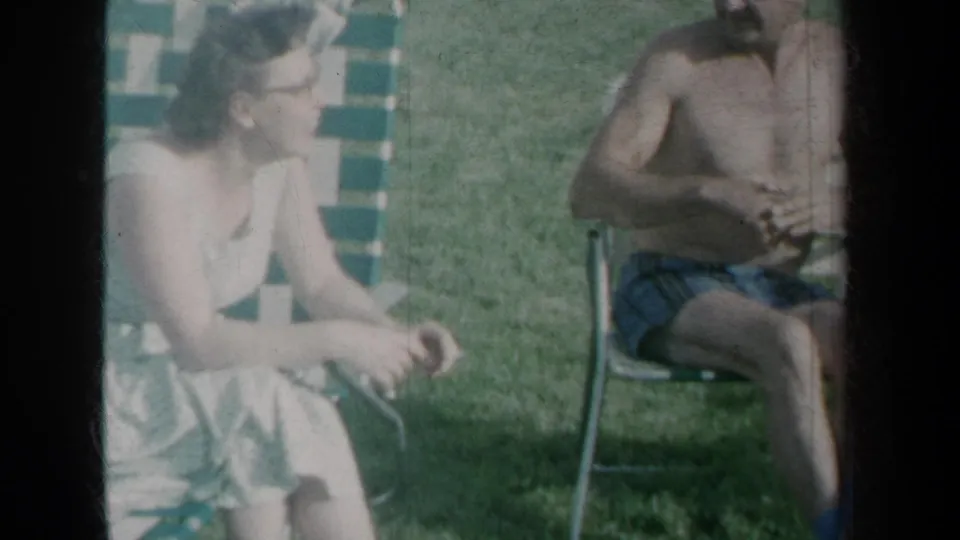 1959 New York home-movie frame with a woman in a light dress seated near a shirtless man on a lawn.