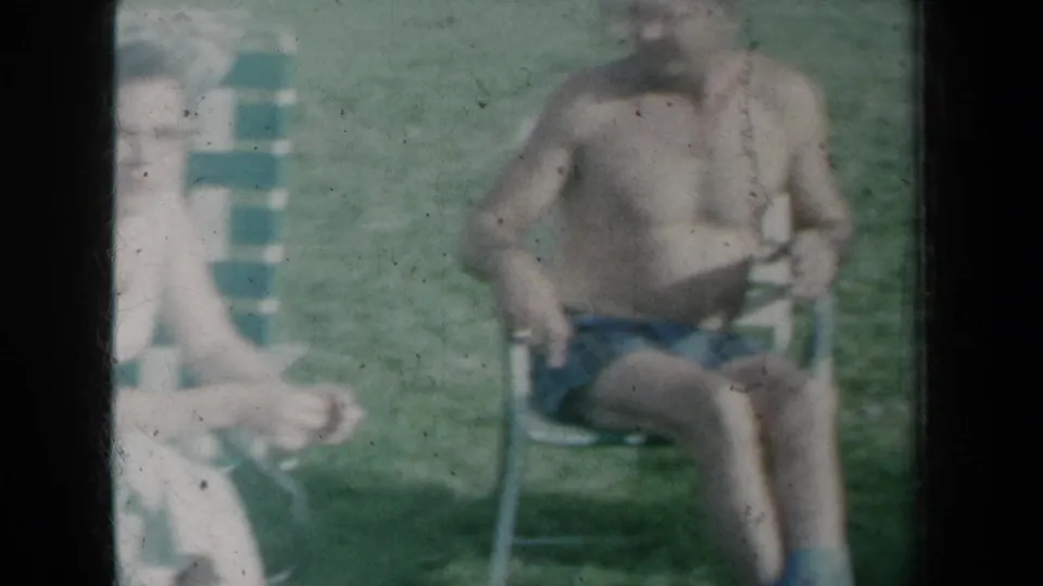 1959 New York frame showing two adults on folding lawn chairs, including a shirtless man in swim trunks.