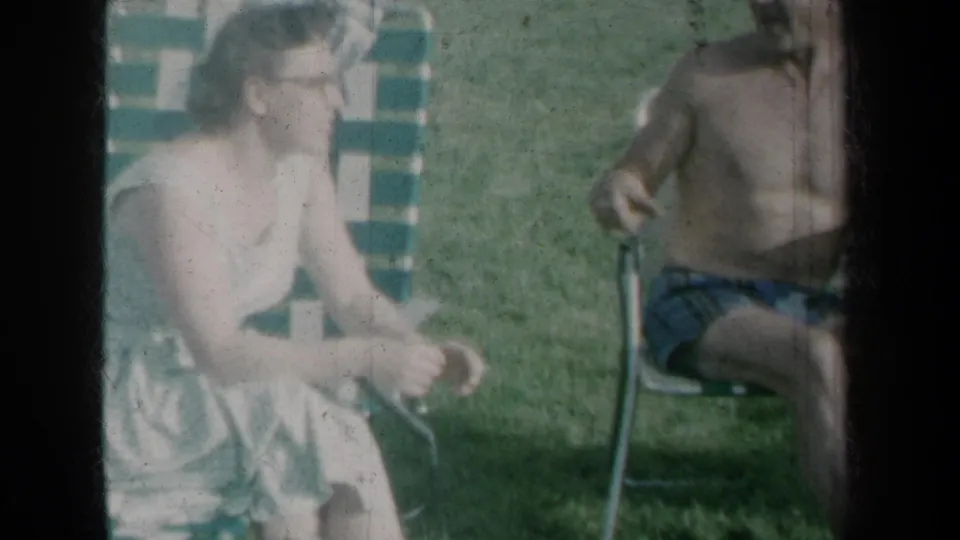 1959 New York home-movie frame of a woman seated on a striped lawn chair in a grassy yard beside another seated adult.