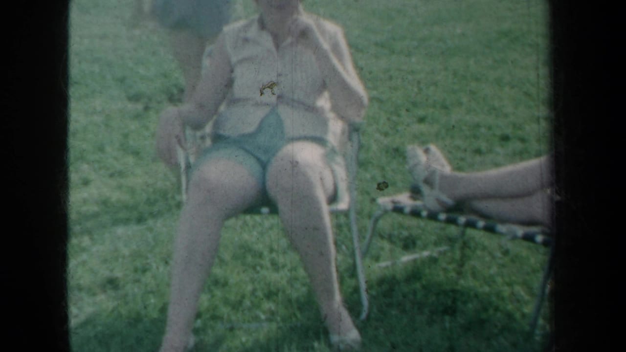 Woman in shorts sits on a metal chair in a grassy yard with another person's legs on a lounge chair.