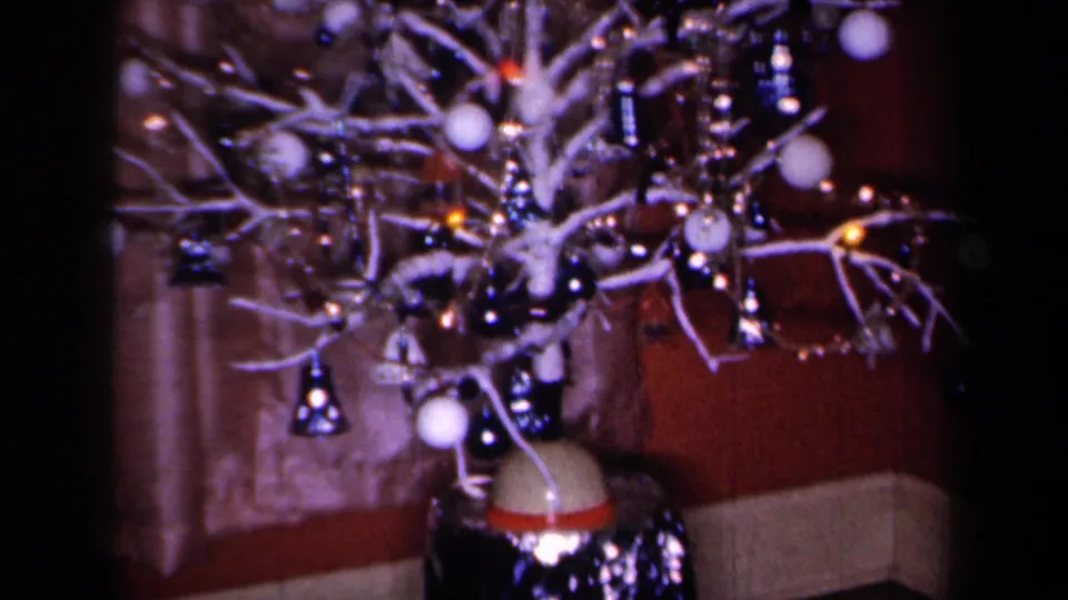 1959 New York home-movie frame of a decorated holiday tree indoors, with ornaments, lights, curtains, and seating visible.