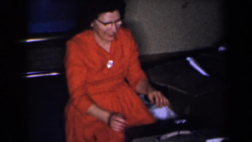1959 New York home interior with an adult woman in a red dress and glasses seated near a suitcase in a dim private room.